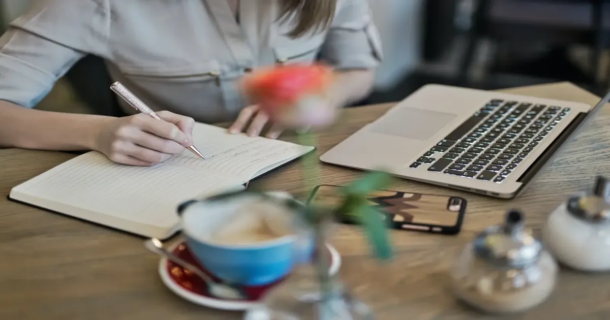 Blogger writing headline ideas in a notebook beside a laptop and coffee